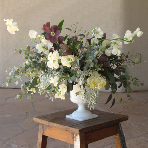 Centerpiece with antique purple hellebores, gunnii eucalyptus, dusty miller, Antique Romantica garden spray roses, and ivory Japanese sweet pea, arranged in a vintage milk glass Grecian urn, by Floral Verde LLC