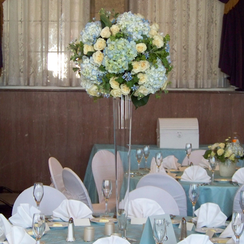 Tall centerpiece with tweedia, pale blue hydrangea, Cream Prophyta roses, bupleurum, and geranium in a 32 pilsner vase