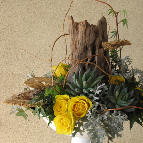 woodland arrangement with a reclaimed wood stump, sponge mushrooms, curly willow tips, yellow roses, Green Trick carnations, ivy,  Pachyveria succulents, and dusty miller in a ceramic pedestal bowl